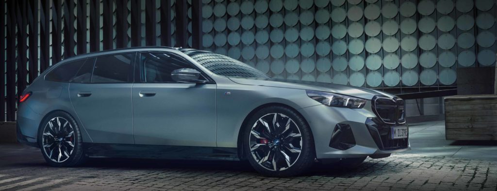 Ein modernes, graues Kombi-Fahrzeug (BMW i5 Touring) steht nachts an einer blauen Ladesäule. Das Auto ist leicht nach links zum Betrachter gewandt, mit eingeschalteten Scheinwerfern, die den dunklen Vordergrund beleuchten. Im Hintergrund befindet sich eine moderne Wand mit kreisförmigen Mustern. Der Gesamteindruck ist dunkel und futuristisch, mit einem Fokus auf Elektromobilität.