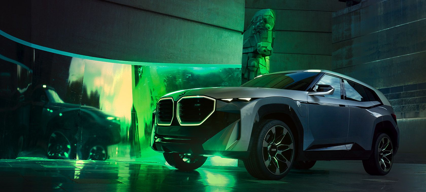 Der kantige und futuristische BMW XM, ein SUV in Grautönen, steht im Fokus vor einem lebendigen grünen Lichtspiel. Eine breite Projektionswand im Hintergrund visualisiert ein weiteres Fahrzeug. Rechts neben dem XM befindet sich eine hohe, dunkle Skulptur, die dem Bild einen Hauch von Kunst und Innovation verleiht.