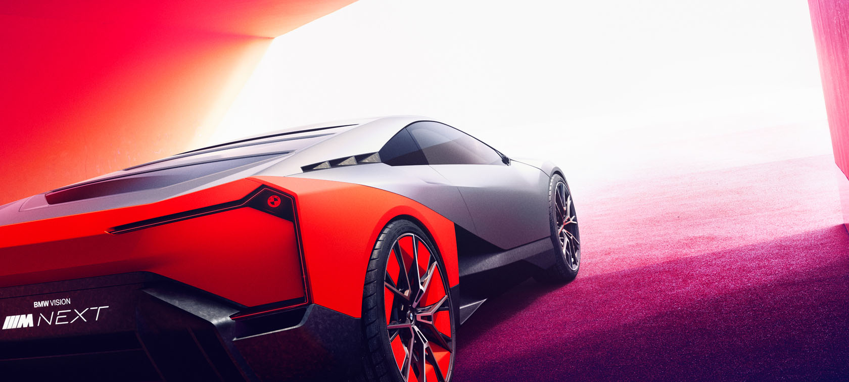 Ein Blick auf das Heck des aufregenden Konzeptfahrzeugs BMW Vision M Next, dessen silberne Karosserie durch leuchtend orangefarbene Elemente ergänzt wird. Die futuristischen Rückleuchten ziehen die Aufmerksamkeit auf sich, ebenso wie die aerodynamisch gestalteten Räder mit roten Akzenten. Die rote und weiße Beleuchtung der Umgebung betont die progressive Formgebung des Sportwagens.