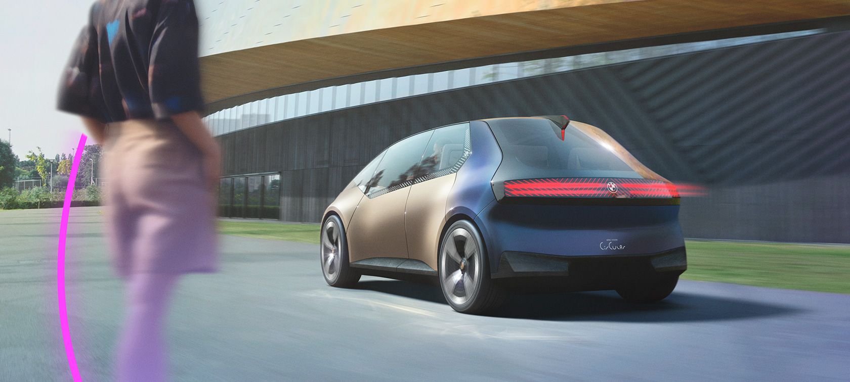 Ein zukunftsweisendes, kleines Fahrzeug in einer auffälligen Kombination aus hellen Braun- und Blautönen, der BMW i Vision Circular, bewegt sich auf einer Straße. Die Heckansicht des Autos präsentiert ein einzigartiges, durchgehendes horizontales Leuchtband als prägnantes Designmerkmal. Im linken Vordergrund ist eine unscharfe Person angedeutet. Die Umgebung im Hintergrund kombiniert moderne Architektur mit natürlichen Elementen wie Bäumen.