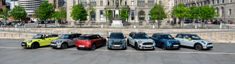 Sechs MINI Cooper Modelle unterschiedlicher Farben stehen nebeneinander auf einem gepflasterten Platz vor einem großen, hellen Gebäude mit vielen Fenstern und klassischer Architektur. Von links nach rechts sind ein gelbes Cabriolet mit offenem Verdeck, ein silberfarbenes Modell mit schwarzen Akzenten, ein rotes Modell, ein hellblaues Modell mit schwarzen Akzenten, ein weißes Modell mit schwarzen Akzenten und ein blaues Modell mit weißen Akzenten zu sehen. Bäume und eine Straße mit geparkten Autos sind im Hintergrund erkennbar.