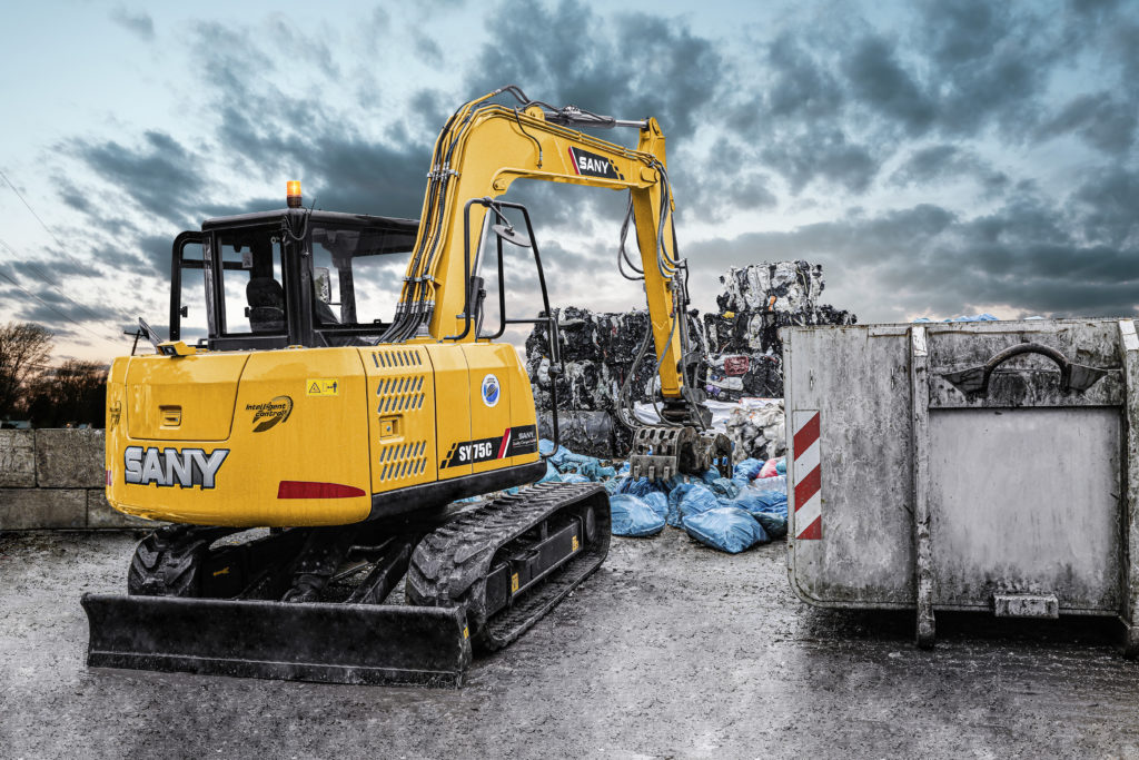 Ein gelber Sany Bagger mit einem Greifer steht auf einem befestigten Untergrund in einem Recyclinghof. Im Hintergrund sind gepresste Wertstoffe in Ballen und ein grauer Container zu sehen. Der Himmel ist bewölkt und düster.