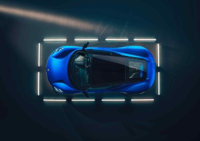 Ein blaues Lotus Emira Sportcoupé mit schwarzem Dach wird von oben in einem dunklen Raum gezeigt. Das Auto ist von rechteckig angeordneten, hellen Leuchtstoffröhren umgeben, die seine Konturen betonen. Die dynamische Form des Fahrzeugs ist aus der Vogelperspektive gut erkennbar.
