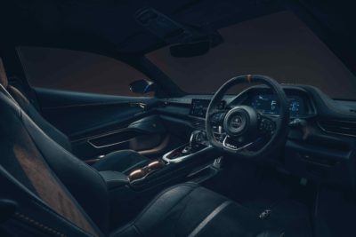 Das dunkle Interieur eines Lotus Emira ist zu sehen. Der Fokus liegt auf dem Fahrerbereich mit dem Sportlenkrad, dem digitalen Kombiinstrument und dem Infotainment-Bildschirm. Die Sitze sind in dunklem Leder mit hellen Akzentstreifen gehalten. Die allgemeine Atmosphäre ist sportlich und hochwertig.