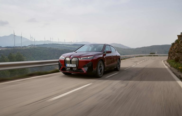 Die Aufnahme zeigt einen roten BMW iX, der auf einer kurvenreichen Straße fährt. Im Hintergrund sind Windräder auf grünen Hügeln zu sehen, was auf eine umweltfreundliche Energieerzeugung hinweist.