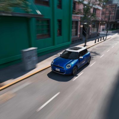 Ein blauer MINI Cooper Electric mit weißem Dach fährt durch eine grüne, städtische Straße aus der Vogelperspektive.