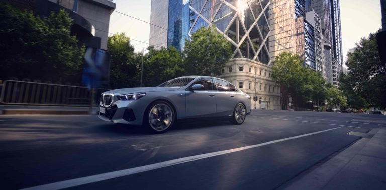 Ein silberner BMW i5 fährt auf einer städtischen Straße. Im Hintergrund sind moderne Hochhäuser und Bäume entlang des Bürgersteigs zu sehen. Die Straße hat weiße Fahrbahnmarkierungen.