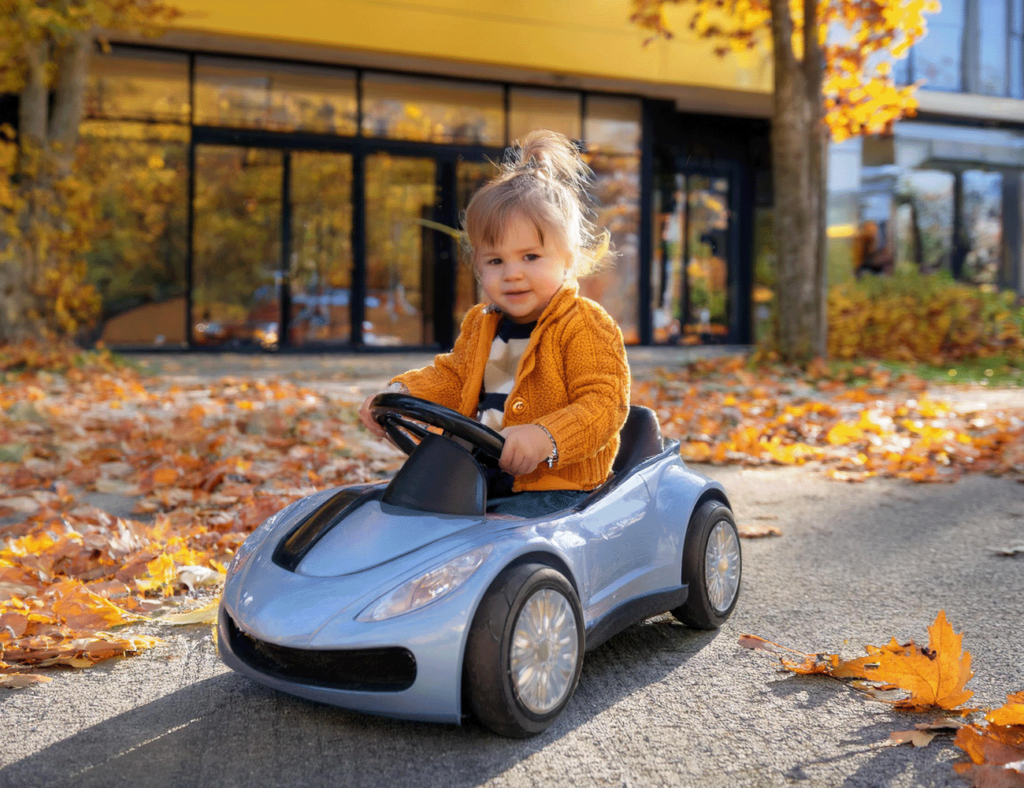 Ein kleines Kind mit blonden Haaren zu einem Dutt gebunden sitzt in einem hellblauen Kinderauto im Freien in Stockach, Baden-Württemberg, Deutschland. Das Kind trägt eine orangefarbene Strickjacke und hält die Hände am Lenkrad. Um das Auto herum liegen bunte Herbstblätter auf dem Boden. Im Hintergrund ist ein Gebäude mit großen Fenstern und herbstlich gefärbten Bäumen zu sehen.