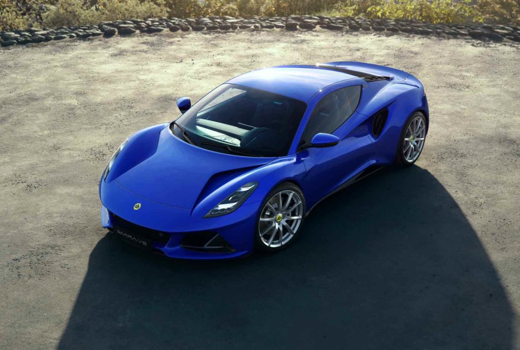Ein blauer Lotus Emira Sportwagen ist von oben schräg vorne auf einem dunklen, asphaltierten Platz abgebildet. Im Hintergrund ist trockene, felsige Landschaft zu sehen.