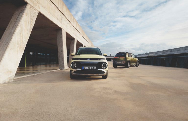 Das Bild zeigt zwei kleine, kastenförmige Hyundai Elektroautos. Das vordere Auto ist hellbeige mit schwarzen Akzenten und hat das Kennzeichen "OF-HY 124E". Es steht frontal zur Kamera. Das hintere Auto ist olivgrün und fährt von der Kamera weg. Die Autos befinden sich auf einer hellgrauen, betonartigen Fläche unter einem hellen Himmel mit einigen Wolken. Links ist ein modernes Gebäude mit großen Betonpfeilern zu sehen.