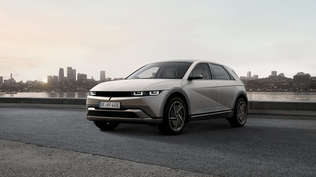 Ein hellgraues, futuristisch anmutendes Elektroauto, ein Hyundai IONIQ 5, steht leicht nach links gedreht auf einer asphaltierten Fläche. Im Hintergrund ist eine Stadtkulisse mit Hochhäusern am Ufer eines Gewässers unter einem grauen Himmel zu sehen. Das Auto hat markante, eckige Formen und helle, rechteckige Scheinwerfer. Das Nummernschild ist gut erkennbar.