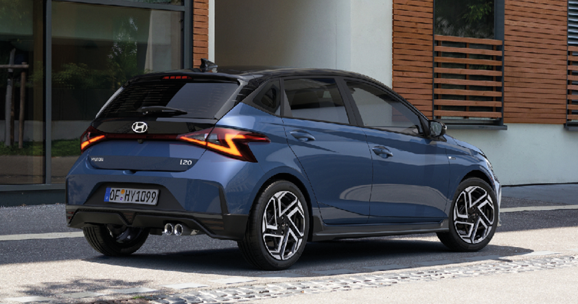 Das Bild zeigt die Rückansicht eines dunkelblauen Hyundai i20. Das Auto steht leicht schräg zur Kamera. Man sieht das Hyundai-Logo, den Schriftzug "i20" und das Kennzeichen "OF-HY 1099". Die Rückleuchten haben eine markante, z-förmige LED-Signatur. Das Auto steht auf einem grauen Untergrund vor einem modernen Gebäude mit großen Fenstern und einer Fassade in Holzoptik.