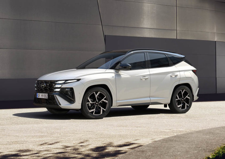 Ein weißer Hyundai Tucson Hybrid steht leicht von vorne links auf einer asphaltierten Fläche in Stockach, Baden-Württemberg, Deutschland. Das Fahrzeug hat ein schwarzes Dach und markante, in den Kühlergrill integrierte Scheinwerfer. Im Hintergrund ist eine moderne Gebäudefassade mit grauen, horizontalen Paneelen zu sehen.