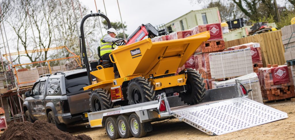 Ein gelber Dumper wird rückwärts eine Rampe hinauf auf einen an einen Pick-up gekoppelten Anhänger gefahren. Ein Arbeiter mit Schutzhelm und Warnweste sitzt am Steuer des Dumpers, im Hintergrund sind Baustellenmaterialien und Gebäude zu sehen.