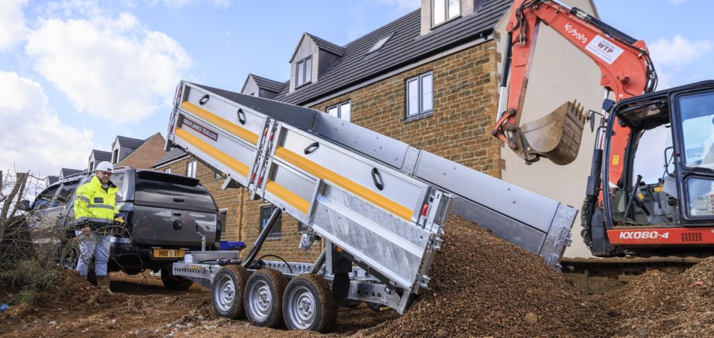 Ein orangefarbener Bagger entlädt Material in einen Brian James Kipper-Anhänger, der an einen dunklen SUV gekoppelt ist. Ein Arbeiter mit Schutzhelm und Warnweste steht neben dem Gespann auf einer Baustelle vor einem Wohnhaus.