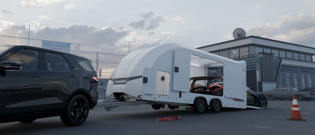 Ein dunkler SUV ist an einen weißen geschlossenen Autotransporter von Brian James Trailers angekoppelt, der ein rotes Sportcoupé transportiert. Das Gespann steht auf einem asphaltierten Platz vor einem Gebäude, das an eine Rennstrecke erinnert, mit Flutlichtmasten im Hintergrund.
