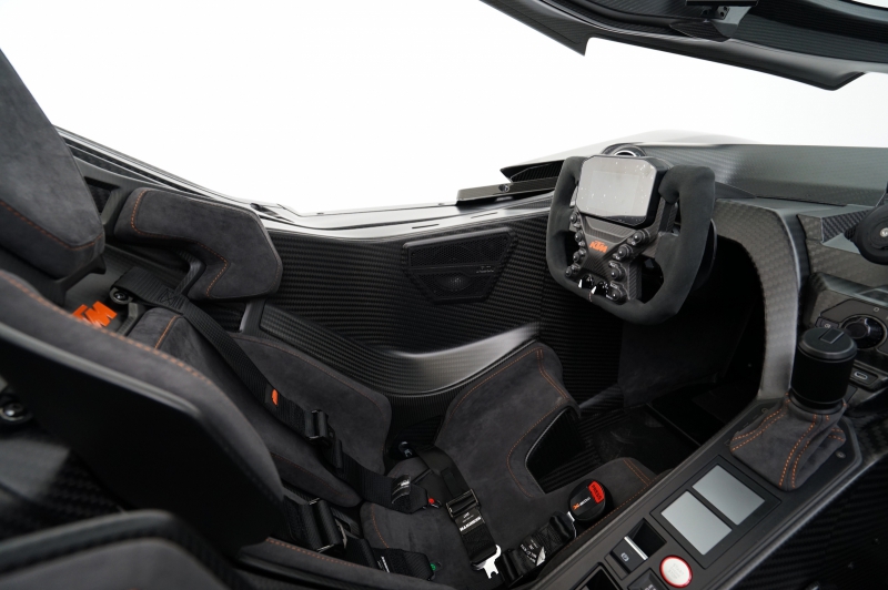 Eine Innenansicht des Fahrerbereichs eines schwarzen KTM X-BOW GT-XR, die das minimalistische, rennsportorientierte Cockpit zeigt. Sichtbar sind das sportliche Lenkrad mit integriertem Display und die stark konturierten Schalensitze mit orangefarbenen Akzenten, umgeben von glänzenden Carbon-Oberflächen.