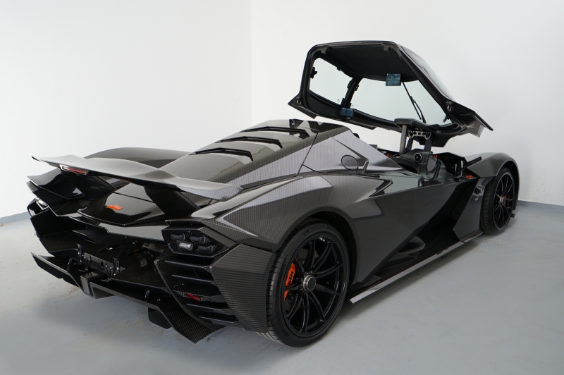 Ein tiefschwarzer KTM X-BOW GT-XR mit offenem Flügeltür-Verdeck steht von hinten links fotografiert vor einer weißen Wand. Die Karosserie zeigt deutliche Carbon-Strukturen und orange Akzente an den Bremssätteln.
