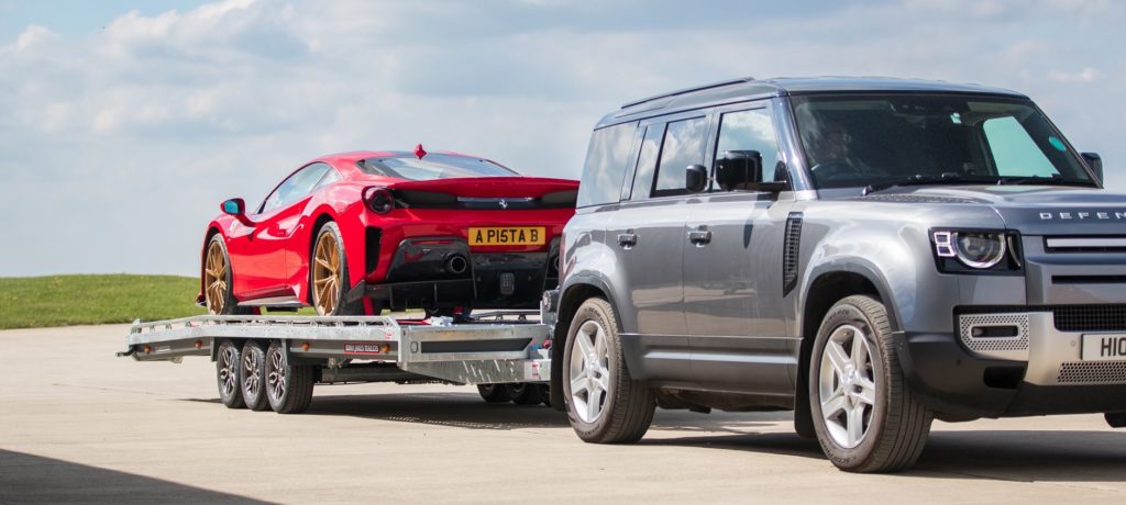 Ein grauer Land Rover Defender zieht einen Anhänger mit einem roten Ferrari, der goldene Felgen besitzt, auf einer asphaltierten Fläche unter leicht bewölktem Himmel. Das Gespann steht seitlich zur Kamera.