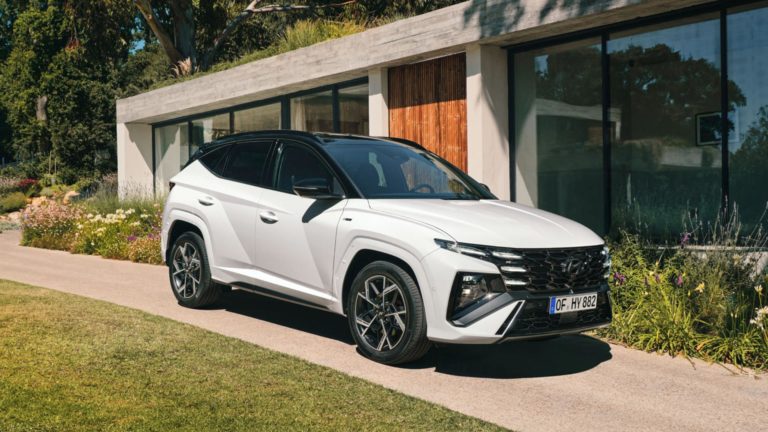 Ein weißer Hyundai Tucson SUV mit schwarzem Dach steht auf einem gepflasterten Weg vor einem modernen Haus mit großen Glasfenstern. Links und rechts des Weges ist gepflegter Rasen und Bepflanzung zu sehen, im Hintergrund Bäume.