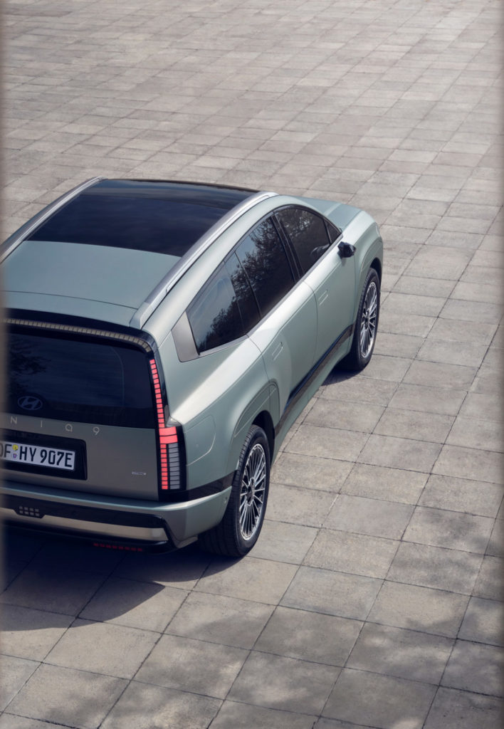 Eine erhöhte Perspektive zeigt das Heck und die Seite eines modernen, hellgrünen SUV, das auf einem gepflasterten Bereich steht. Das Fahrzeug hat ein dunkles Panoramadach und markante vertikale Rückleuchten. Das Kennzeichen "OF HY 907E" ist sichtbar. Die Aufnahme wird von dunklen, unscharfen Elementen im Vordergrund gerahmt.