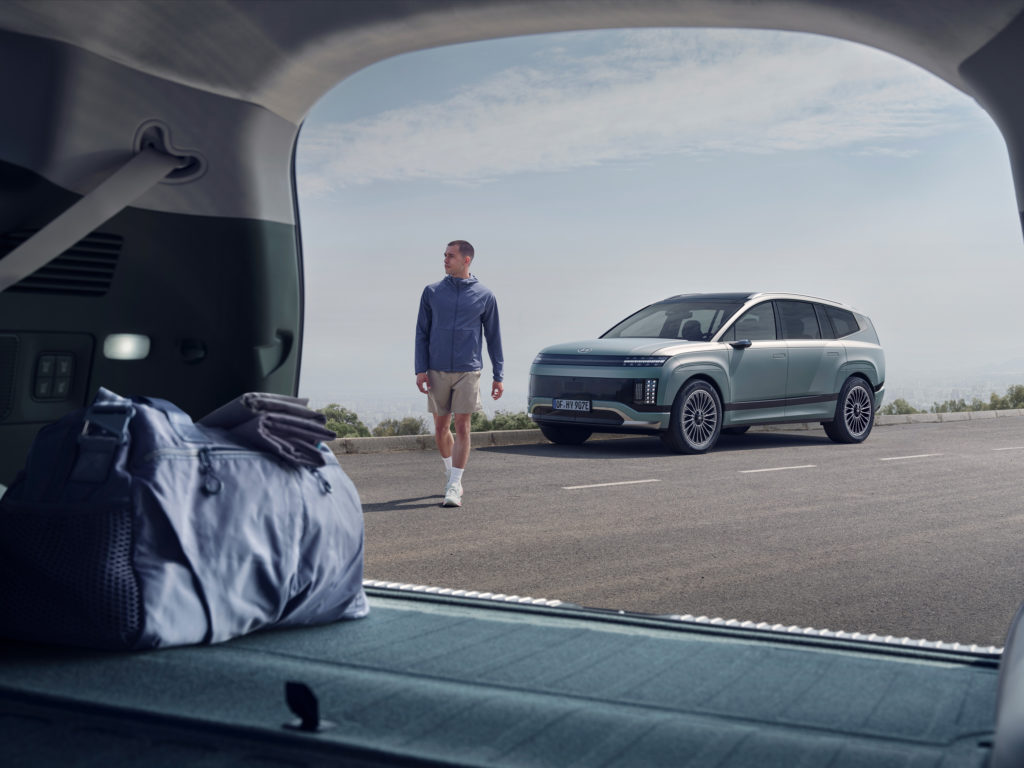 Blick aus dem geöffneten Kofferraum eines modernen SUVs auf einen Mann in Sportkleidung, der auf einer Straße am Meer auf das Fahrzeug zuläuft. Im Vordergrund sind eine Reisetasche und Handtücher im Kofferraum zu sehen. Das hellgrüne SUV steht auf der rechten Seite der Straße, während der Mann von links kommt. Der Himmel ist leicht bewölkt.