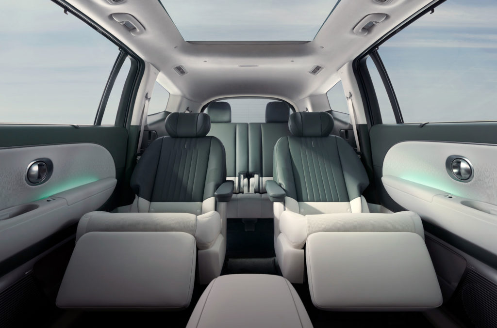 Blick in den geräumigen Innenraum eines modernen Autos mit luxuriösen, verstellbaren Sitzen. Die beiden Vordersitze sind in einer entspannten Position mit hochgeklappten Beinauflagen dargestellt. Im Hintergrund sind weitere Sitzreihen zu sehen. Der Innenraum ist in hellen und dunklen Grautönen gehalten, ergänzt durch ambiente Beleuchtung in den Türverkleidungen und ein großes Panorama-Glasdach, das für viel Licht sorgt.
