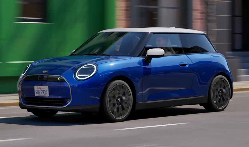 Ein blauer Mini Cooper SE, von vorne schräg links aufgenommen, fährt auf einer Straße. Das Fahrzeug hat ein hellgraues Dach und runde Scheinwerfer mit einem LED-Ring. Im Hintergrund sind unscharfe Gebäude in Grün- und Brauntönen zu sehen, was Bewegung suggeriert.