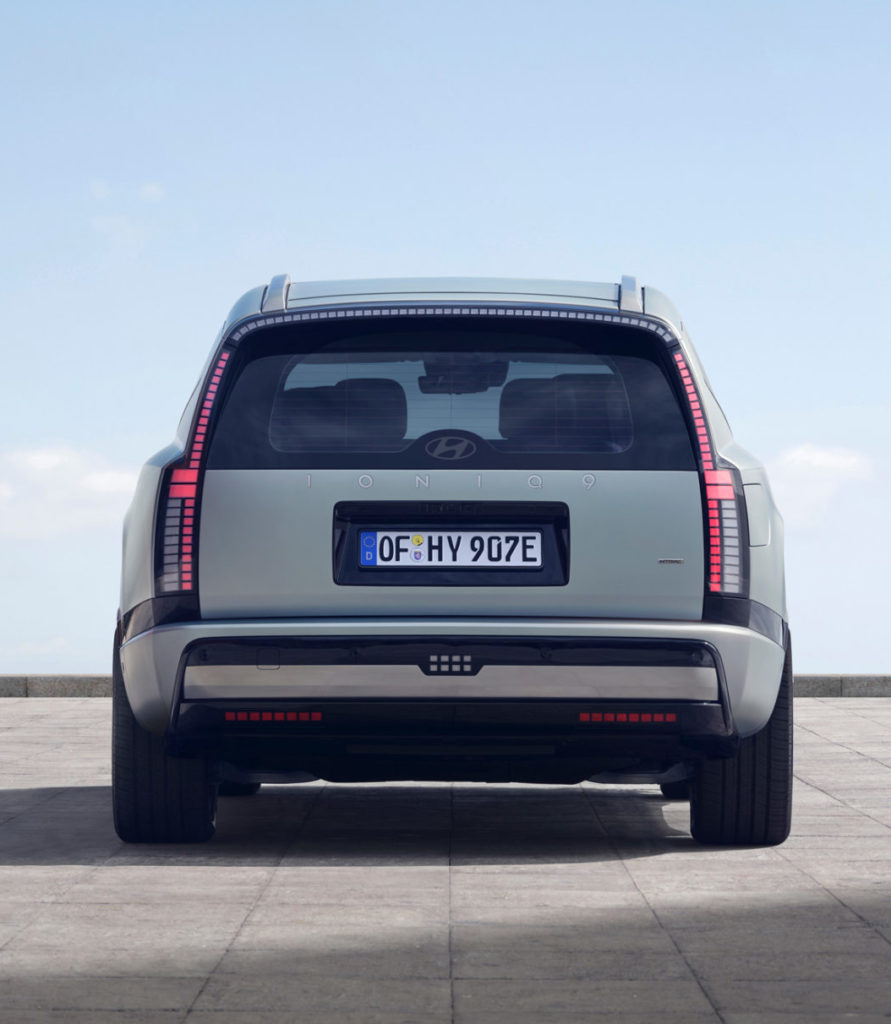 Rückansicht eines modernen, hellgrauen oder hellgrünen SUVs, das auf einer weiten, hellen Asphaltfläche unter einem weitläufigen blauen Himmel parkt. Das Fahrzeug zeigt seine markanten vertikalen Rückleuchten und das Kennzeichen "OF HY 907E". Es ist zentral im Bild positioniert, mit viel Freiraum um es herum, was seine Größe und Präsenz unterstreicht.