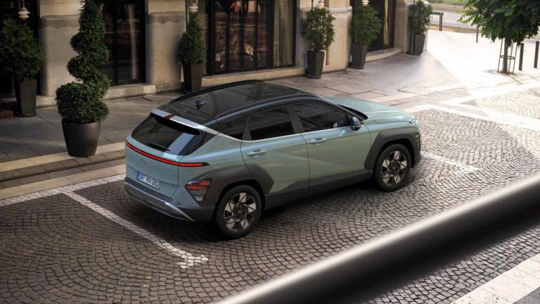 Eine erhöhte Draufsicht auf das Heck eines mintgrünen Hyundai Kona SUV, der auf einem gepflasterten Gehweg vor einem Geschäft mit Schaufenstern und Pflanzen in Töpfen parkt. Das Fahrzeug hat ein schwarzes Dach und schwarze Radkästen, die seine robuste Erscheinung unterstreichen.