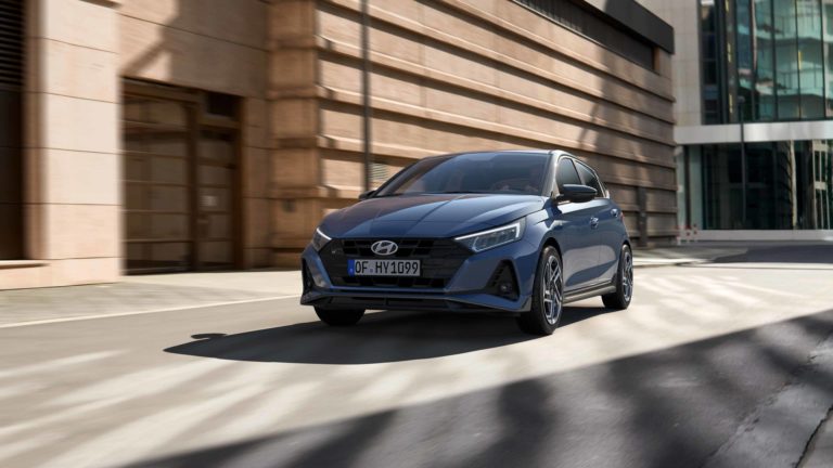 Ein blauer Hyundai i20 fährt schnell auf einer städtischen Straße. Die Umgebung mit modernen Gebäuden ist durch die Geschwindigkeit leicht verschwommen.
