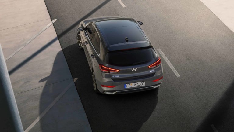 Eine erhöhte Draufsicht auf das Heck eines grauen Hyundai i30, der auf einer Straße mit Markierungen steht. Das Fahrzeug hat ein dunkles Panoramadach und ist dynamisch in Szene gesetzt.