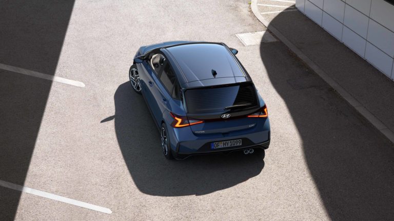 Eine erhöhte Draufsicht auf das Heck eines blauen Hyundai i20, der auf einem asphaltierten Parkplatz steht. Das Fahrzeug hat ein sportliches Design mit auffälligen Rückleuchten.