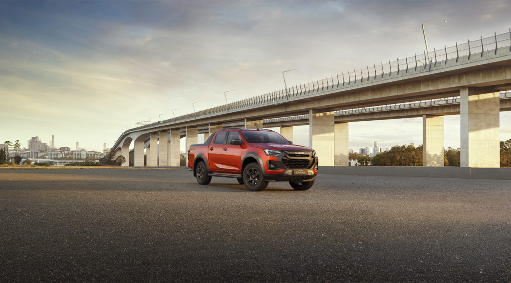Frontansicht eines roten Isuzu D-MAX Pickup-Trucks, der auf einem asphaltierten Platz unter einer Autobahnbrücke steht, mit einer Stadtkulisse im Hintergrund.