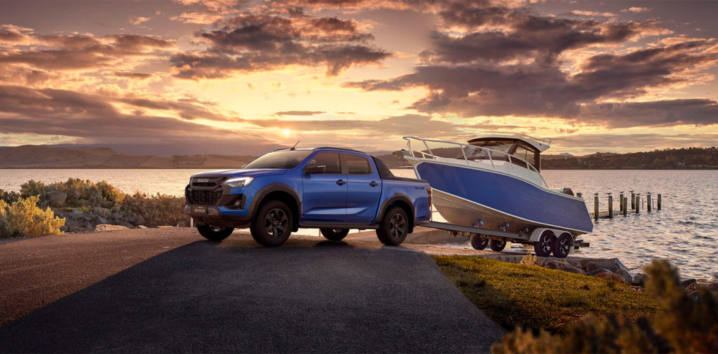 Ein blauer Isuzu D-MAX Pickup-Truck zieht einen Bootsanhänger mit einem weißen Boot. Das Gespann befindet sich an einem Seeufer bei Sonnenuntergang.