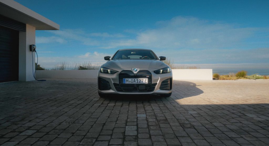 Frontalansicht eines grauen BMW i4 Gran Coupé, das auf einem gepflasterten Vorplatz vor einer modernen Villa und mit Meerblick im Hintergrund steht. Das Fahrzeug ist an eine Ladesäule angeschlossen.
