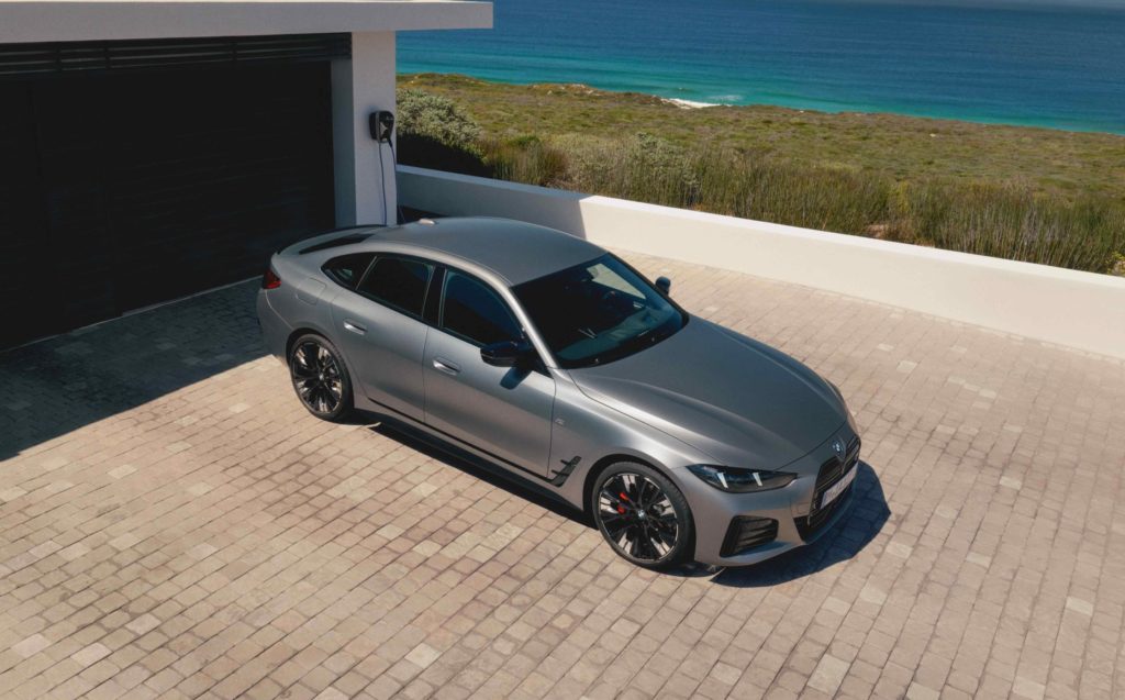 Eine erhöhte Ansicht auf ein graues BMW i4 Gran Coupé, das vor einer modernen Villa mit Garage und Meerblick parkt und an eine Ladesäule angeschlossen ist. Die Szene ist von hellem Sonnenlicht beleuchtet.