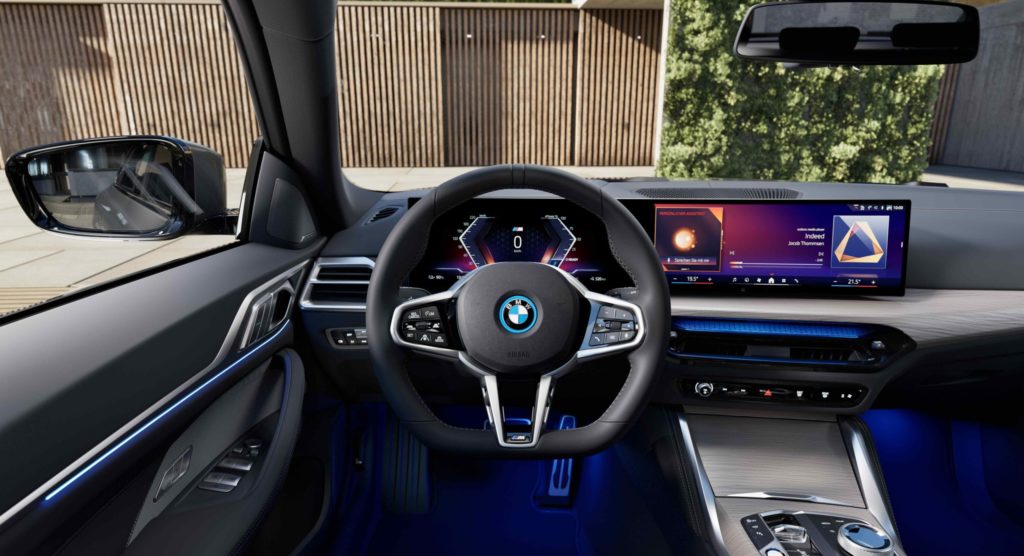 Innenansicht eines modernen BMW i4, das Armaturenbrett ist mit einem breiten digitalen Display ausgestattet, das Lenkrad und der Sitz haben sportliche Details. Die Ambiente-Beleuchtung in Blau schafft eine futuristische Atmosphäre.