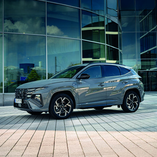 Ein grauer Hyundai Tucson steht seitlich auf einem gepflasterten Boden vor einem modernen Gebäude mit Glasfassade.