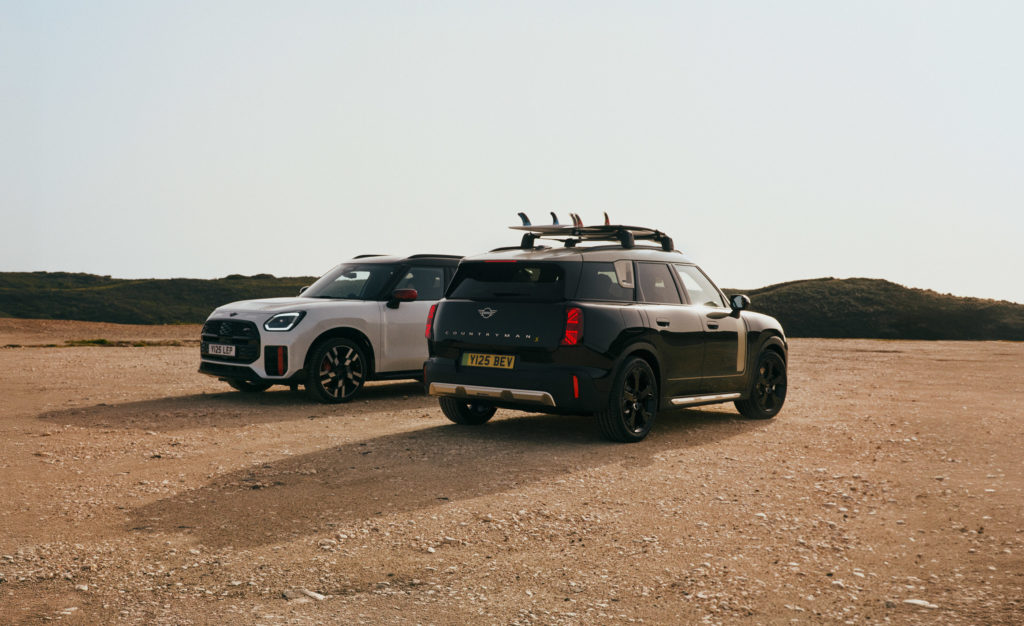 Zwei MINI Countryman stehen nebeneinander in einer sandigen Landschaft. Der vordere, schwarze Wagen hat Surfbretter auf dem Dach, der hintere ist weiß.