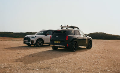 Zwei MINI Countryman stehen nebeneinander in einer sandigen Landschaft. Der vordere, schwarze Wagen hat Surfbretter auf dem Dach, der hintere ist weiß.