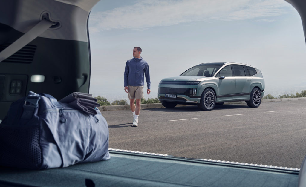 Ein hellgrauer Hyundai steht auf einer Straße. Ein Mann in Sportkleidung geht auf das Fahrzeug zu. Das Foto ist aus dem geöffneten Kofferraum eines anderen Autos gemacht, in dem eine Sporttasche liegt.