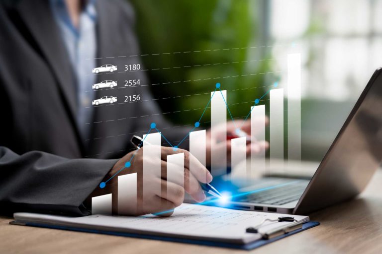 Eine Person in Anzug arbeitet am Laptop mit einem Notizblock, während eine digitale Grafik mit Autos und Daten eingeblendet ist.