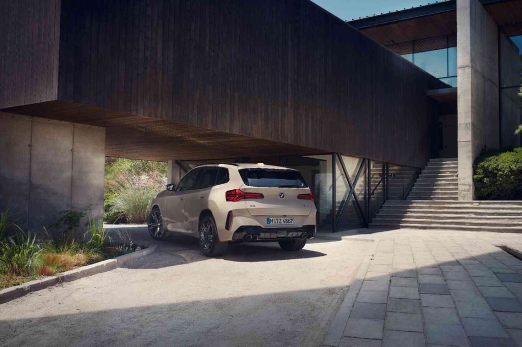 Ein beigefarbener BMW X3 M50 xDrive steht auf einer gepflasterten Auffahrt und steht unter dem Holzvordach eines modernen Hauses. Das Heck und die sportlichen Endrohre sind deutlich zu sehen.