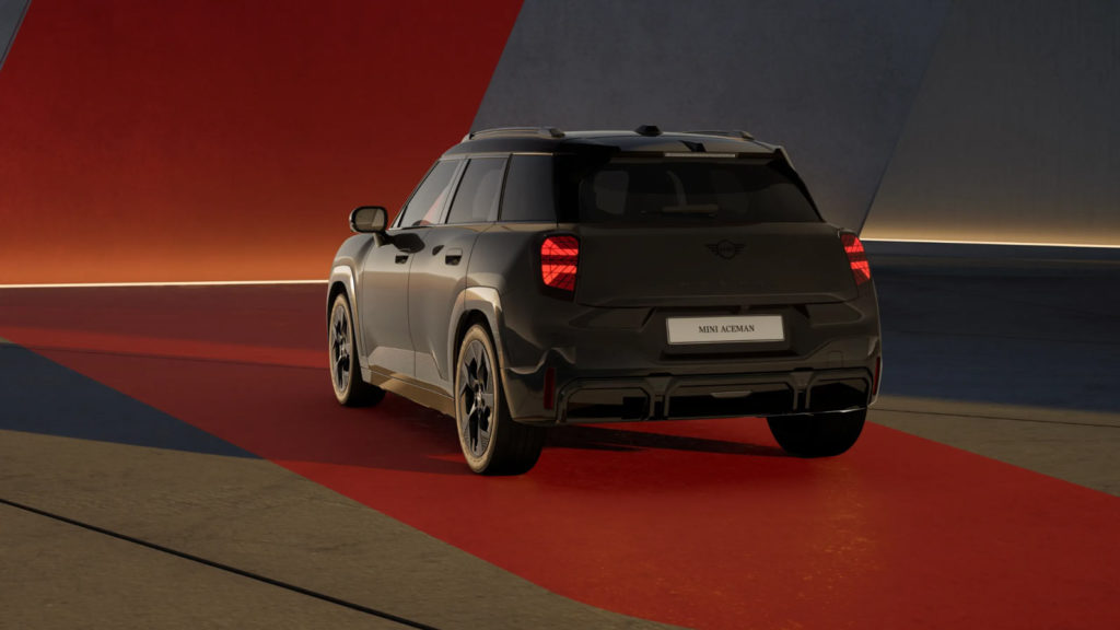 Der schwarze MINI Aceman E John Cooper Works Trim ist von hinten rechts auf einem roten Streifen zu sehen. Die Rückleuchten leuchten, und die Umgebung ist in schummriges Licht getaucht mit roten und grauen Wänden im Hintergrund.