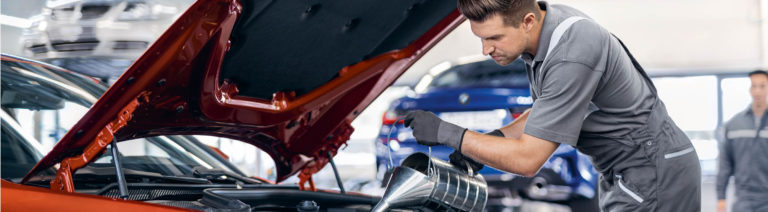 Ein Mechaniker füllt Öl in einen roten BMW mit geöffneter Motorhaube. Im Hintergrund sind weitere Fahrzeuge in einer hellen Werkstatt zu sehen.