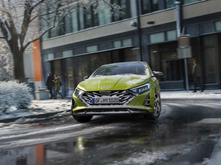 Ein hellgrüner Hyundai i20 fährt durch eine verschneite Stadtstraße mit Schneeresten auf dem Auto und Boden.