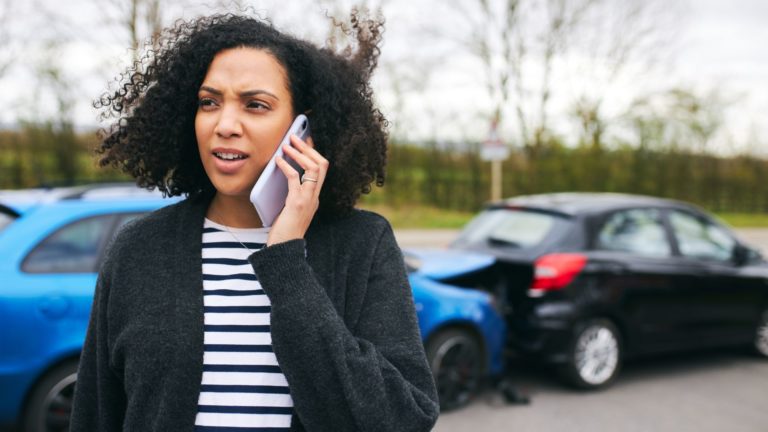 Besorgte Frau telefoniert nach einem Auffahrunfall; im Hintergrund stehen ein blauer und ein schwarzer Pkw.