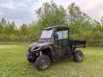 Grünes Diesel-UTV mit geschlossener Kabine und offener Ladefläche auf einer Wiese vor Bäumen und bewölktem Himmel.