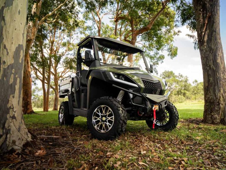 Ein olivgrünes Diesel-UTV mit offener Kabine und Ladefläche steht im Schatten hoher Bäume auf einer bewaldeten Grünfläche.